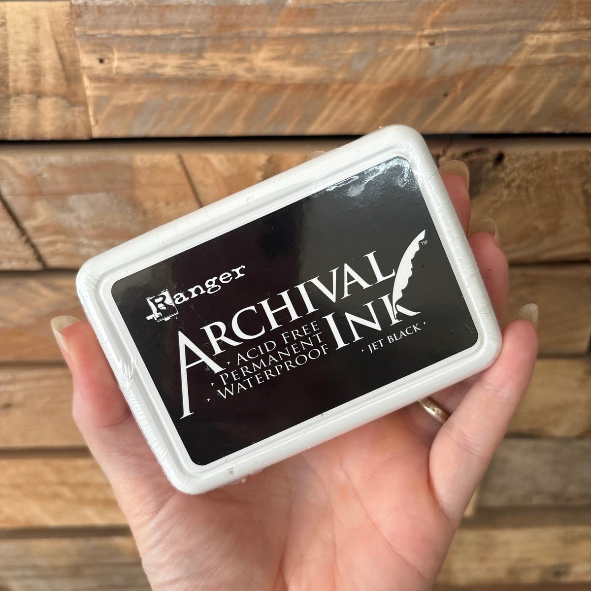 A hand holding a square Archival Ink pad in a white plastic case against a rustic wooden plank background.