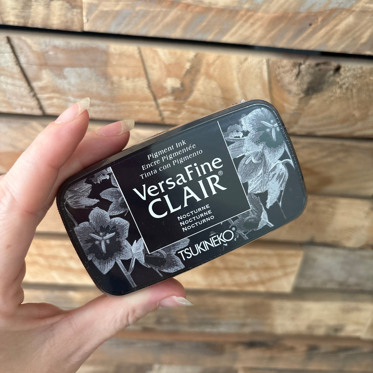 A hand holding a black rectangular tin of VersaFine Clair against a rustic wooden plank background.