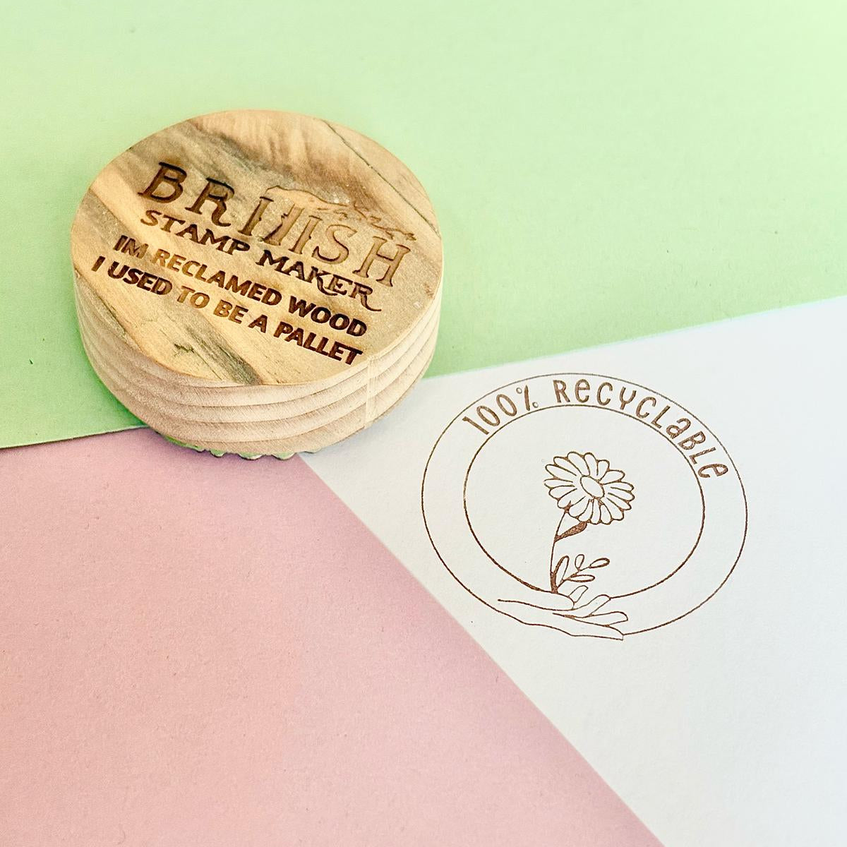 Reclaimed wood round block with British stamp maker logo engraved, on a green and pink background with a vintage sepia versafine ink stamped on white. the stamp design is 100% recyclable with hands holding a flower in a circle.