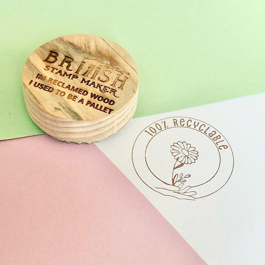Reclaimed wood round block with British stamp maker logo engraved, on a green and pink background with a vintage sepia versafine ink stamped on white. the stamp design is 100% recyclable with hands holding a flower in a circle.