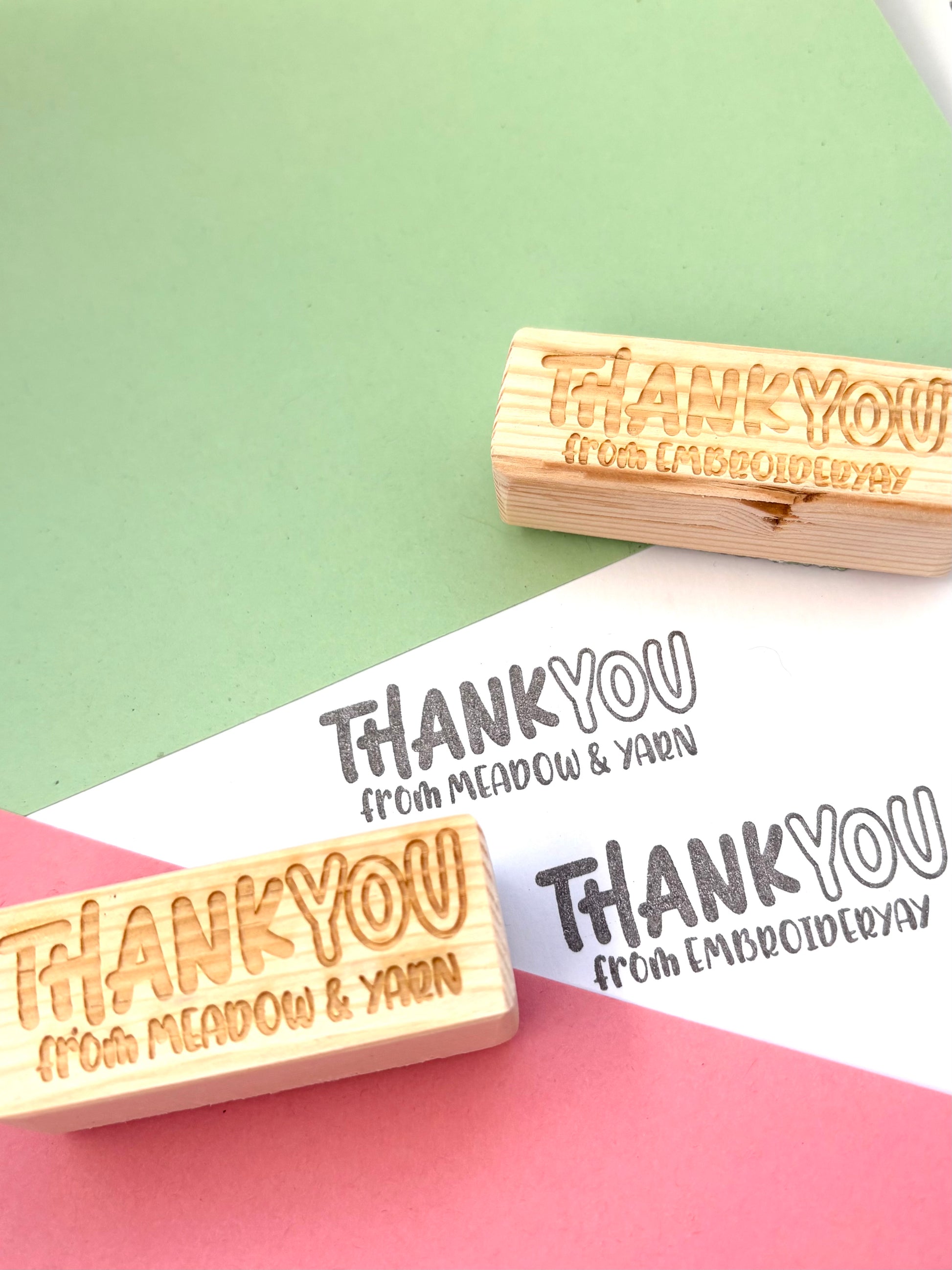 Personalised thank you stamp with thank you text in bold on the top and from your business name on the bottom in smaller text. On a pink and green background with reclaimed wood block handles. 