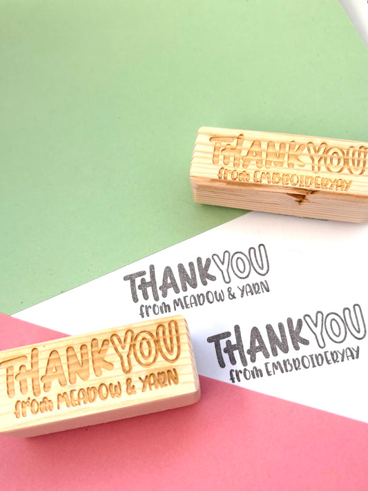 Personalised thank you stamp with thank you text in bold on the top and from your business name on the bottom in smaller text. On a pink and green background with reclaimed wood block handles. 