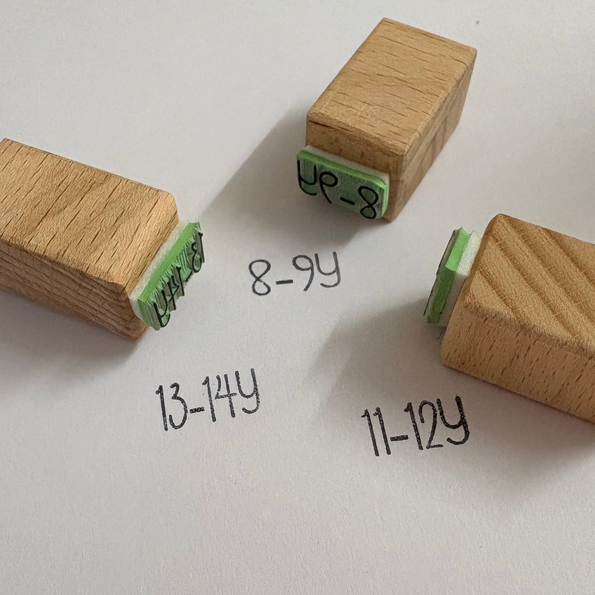 Four wooden block stamps with green ink pads are arranged on a white sheet of paper, with handwritten age labels ('8-9Y', '13-14Y', '11-12Y', and '8-9Y') near them.