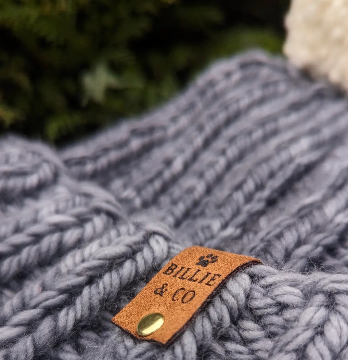 Brown faux suede label on a blue crochet bobble hat. Featuring billie and co logo with a paw print  and a gold rivet fastening.