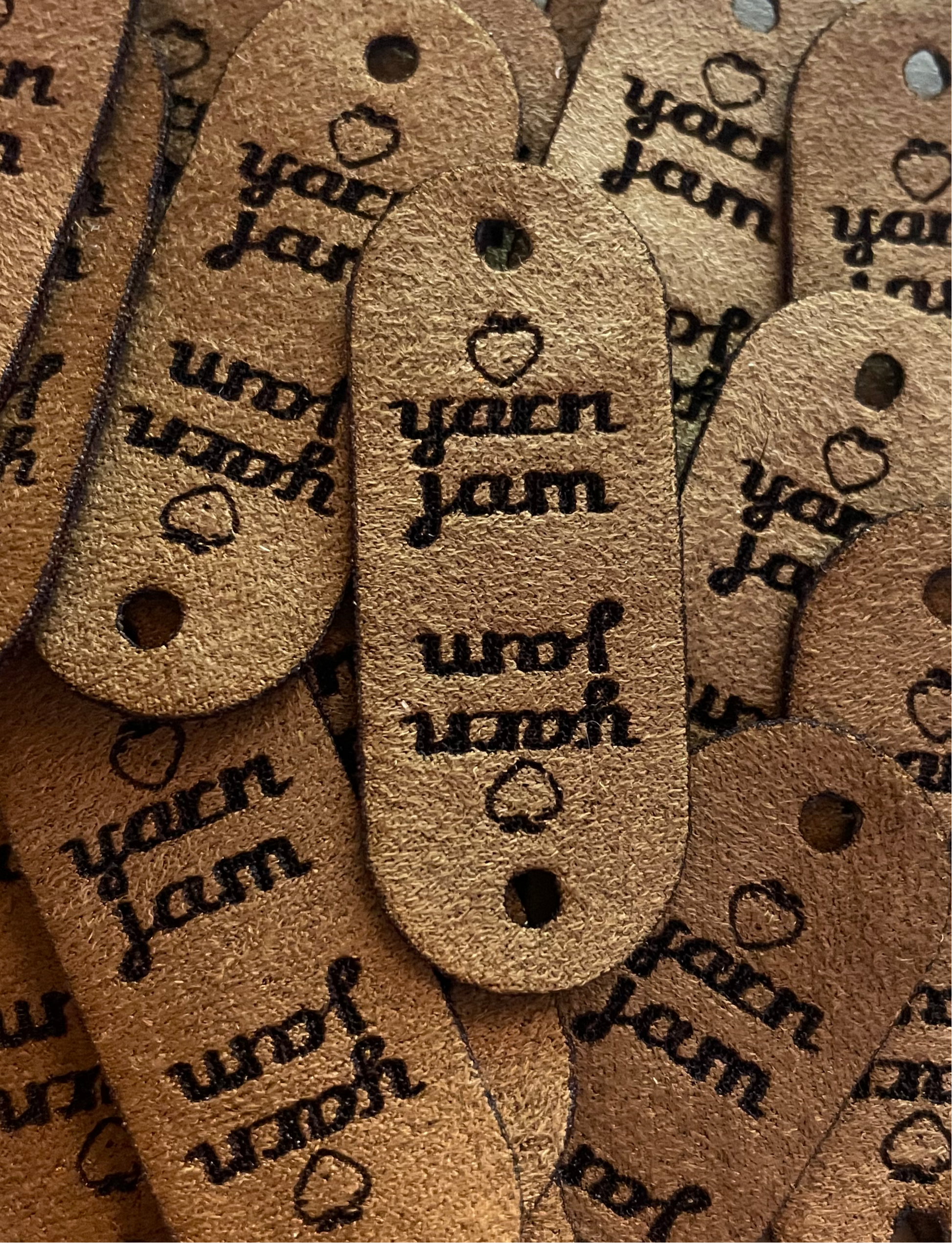 Brown faux suede labels with rounded edges. One hole for a rivet  fastener. Featuring yarn jam logo including a small strawberry.
