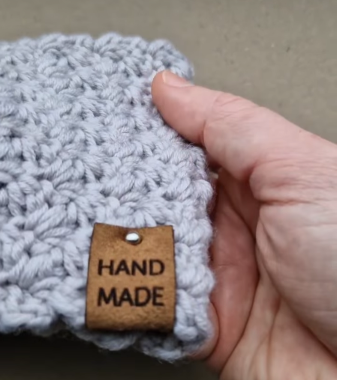 Brown faux suede label with hand made text, attached to a grey crochet headband with a silver rivet fastener.