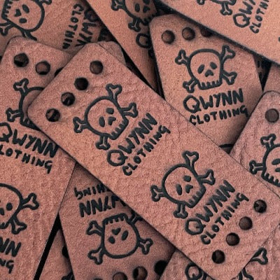 Qwynn Clothing logo on a brown faux leather fold over tag with four holes to hand sew into garments. The logo includes a skull and cross bones.