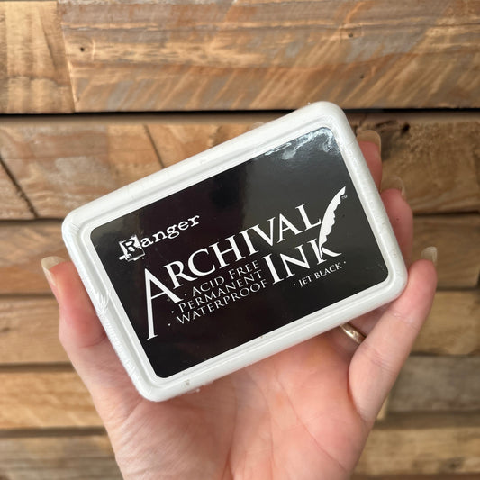 A hand holding a square Archival Ink pad in a white plastic case against a rustic wooden plank background.