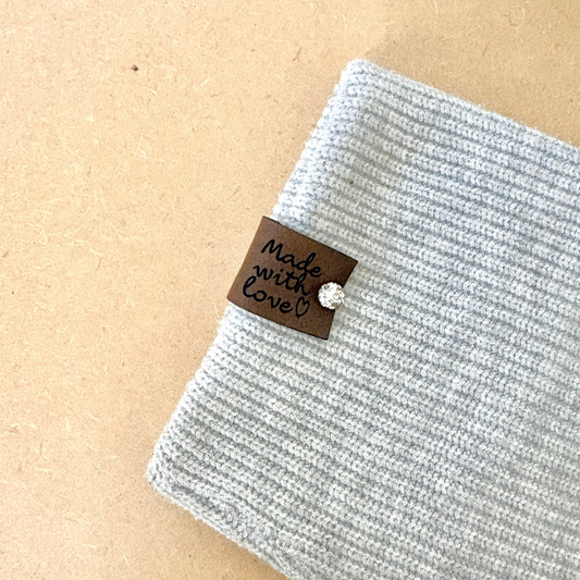 A close-up of a light gray ribbed knit fabric item, likely a beanie or folded textile, placed on a tan kraft-paper background. A small brown fabric tag with handwritten text reading 'Made with Love' and a clear rhinestone pin are attached.