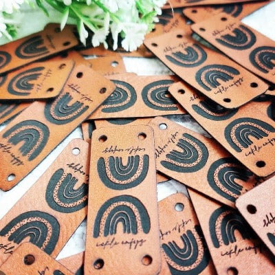 ickle enfys rainbow logo on brown faux leather tags with two holes for hand sewing onto handmade clothing.
