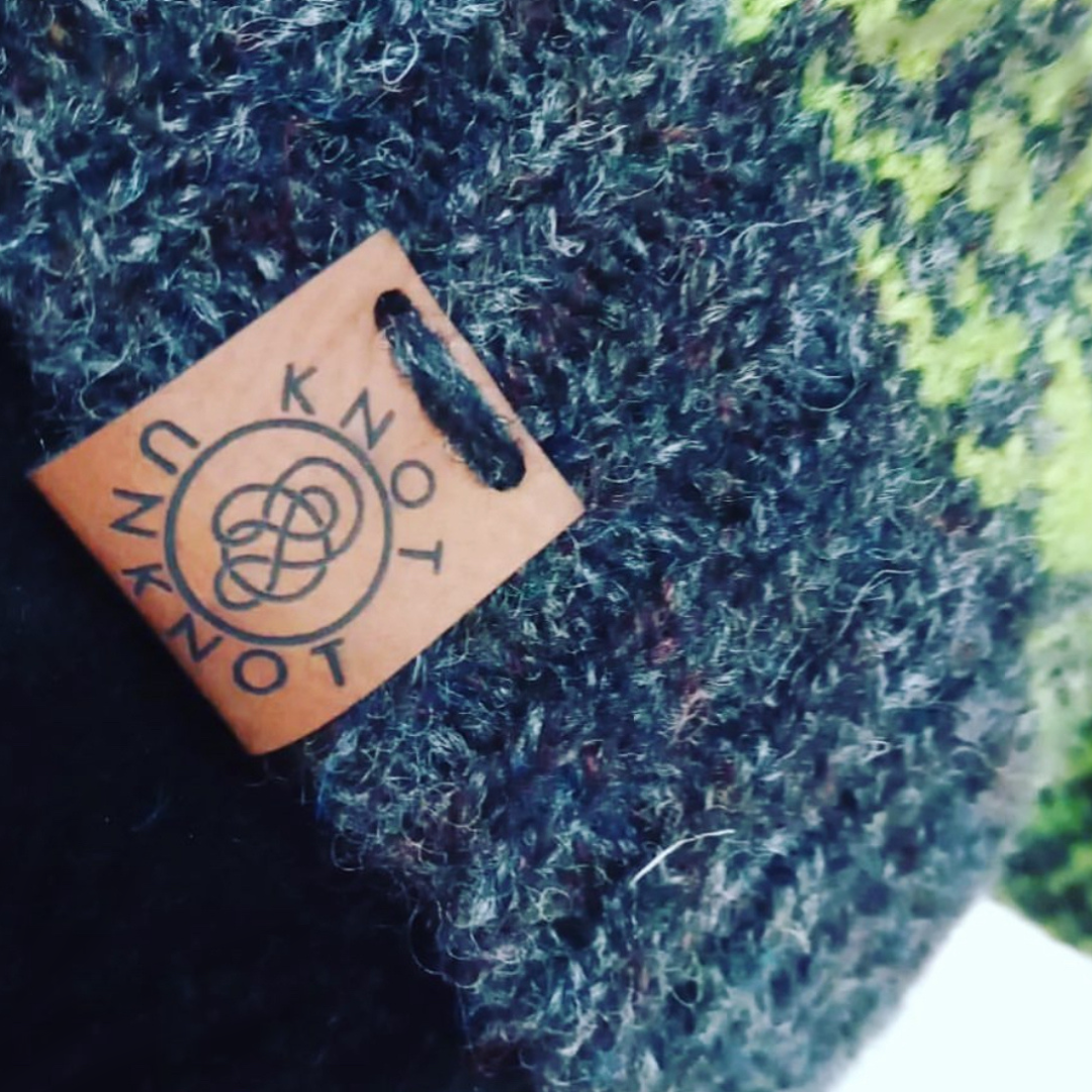knot unknot logo on a brown faux leather tag hand sewn onto a handmade hat made from blue and green wool.