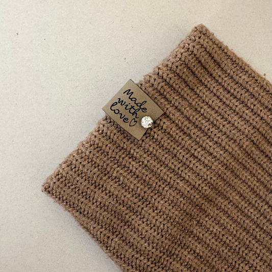 A close-up of a ribbed knit brown fabric item, likely a scarf or sleeve cuff, laid on a light beige background. A small sewn label reads 'made with love?' with a tiny round metallic charm.